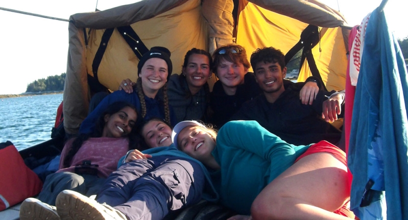 A group of people sit under a tarp shelter set up on a sailboat. 